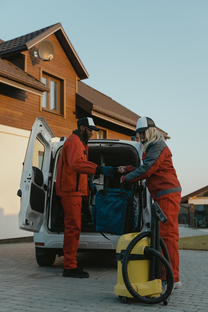 Two professional cleaners in work uniforms unloading equipment from a van in a residential area.